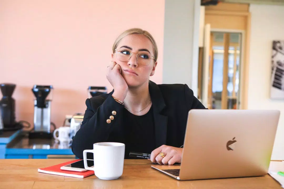 Mujer con gafas sentada en una mesa de café, aburrida o pensativa, con una computadora portátil abierta y una taza de café a su lado.