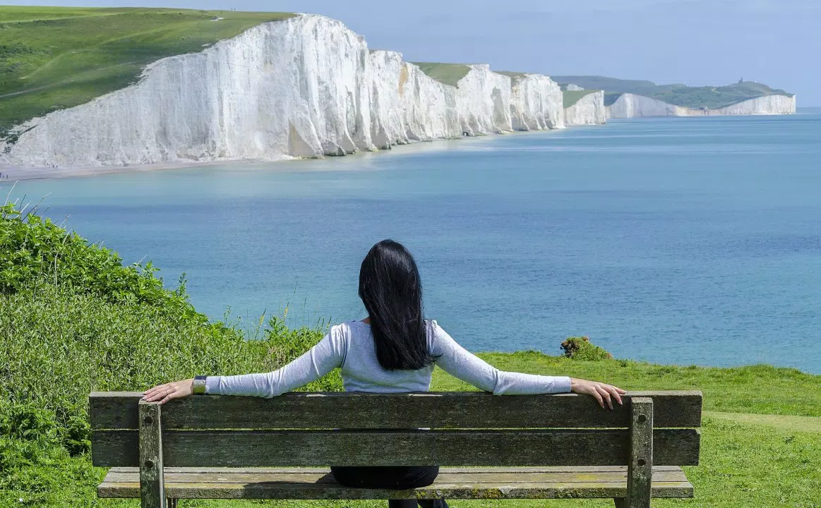 Una mujer sentada en un banco con vistas a un tranquilo mar azul y espectaculares acantilados blancos bajo un cielo despejado.