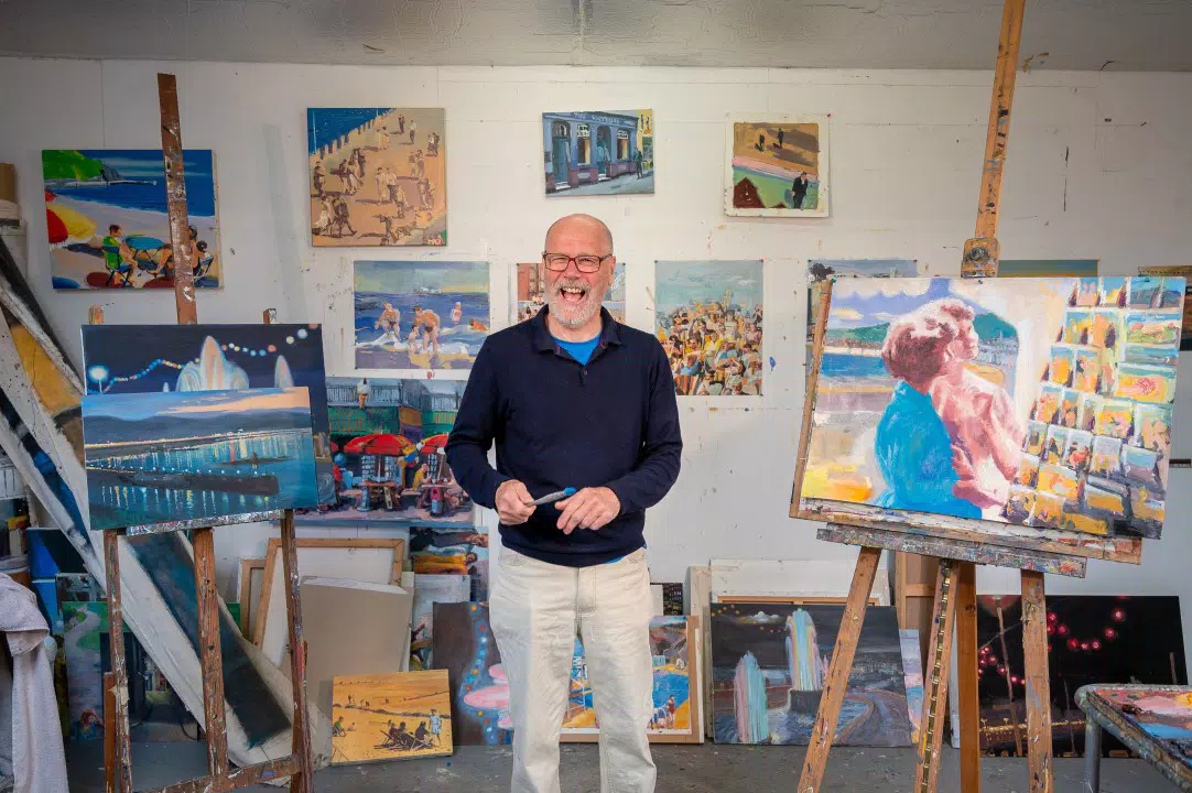 Un hombre sonriente se encuentra en un estudio de arte rodeado de coloridas pinturas sobre caballetes.