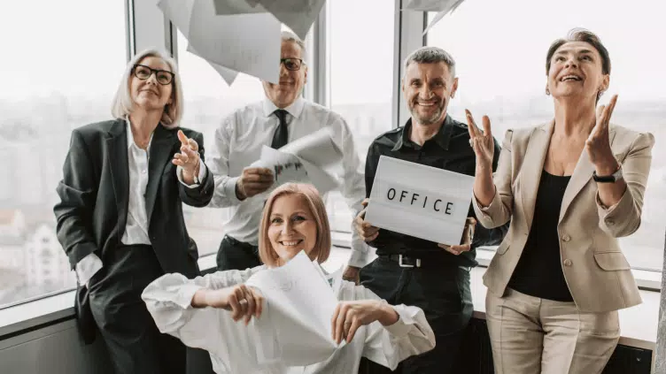 Grupo de cinco compañeros de oficina sonriendo, lanzando papeles al aire, celebrando con un cartel que dice "oficina".