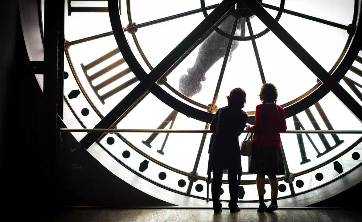 Dos personas mirando por una gran ventana redonda con forma de reloj, recortadas contra un cielo brillante.