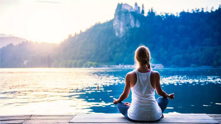 Una mujer practica yoga en posición de loto en un muelle de madera con vistas a un lago sereno con una montaña boscosa al fondo al atardecer.