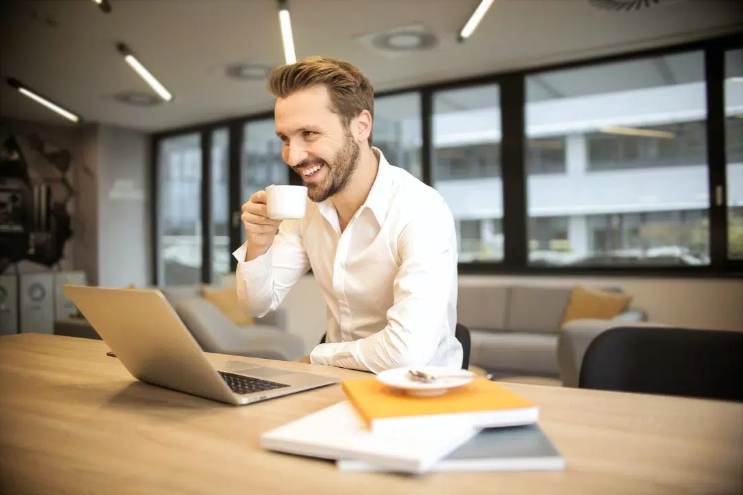 Un hombre vestido con un traje informal de negocios sonriendo y tomando café, mientras usa una computadora portátil en un escritorio de oficina moderno.