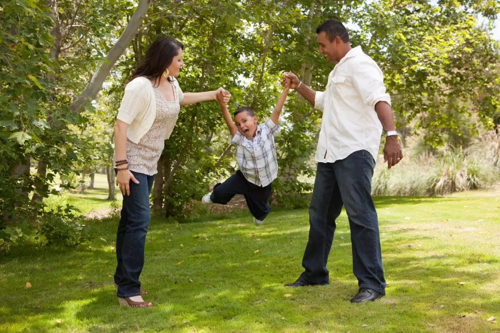 Una familia alegre jugando al aire libre, con los padres levantando a su hijo de los brazos mientras éste se balancea entre ellos en un parque con césped, lo que ilustra un momento de bienestar y conexión.