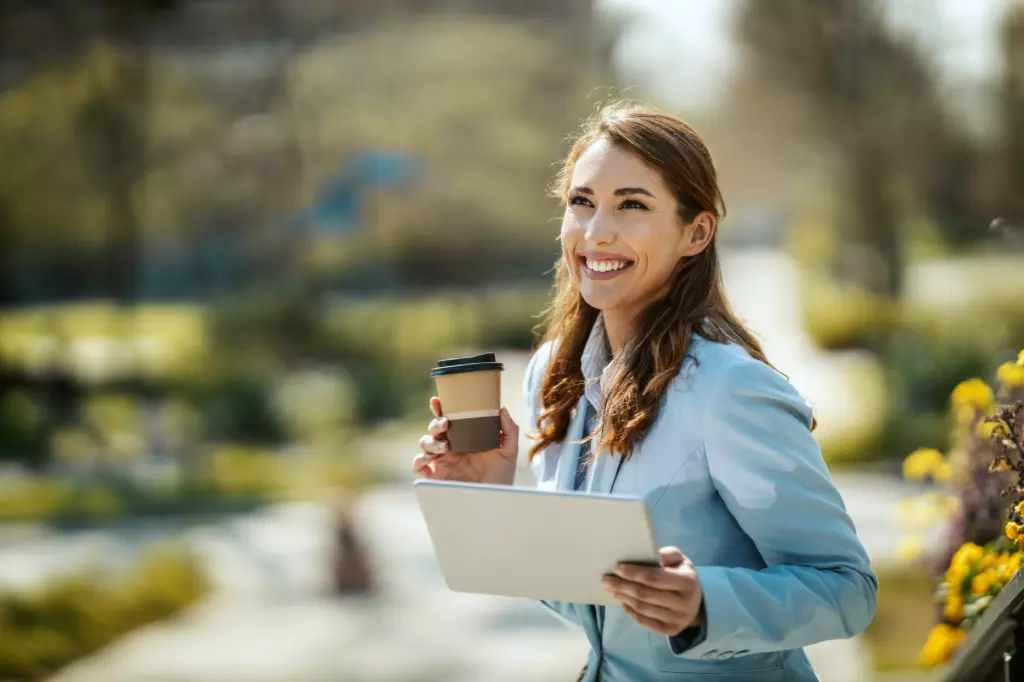 Una mujer sonriente sosteniendo una taza de café y una tableta mientras está de pie al aire libre en un parque soleado, luciendo como el epítome del bienestar.