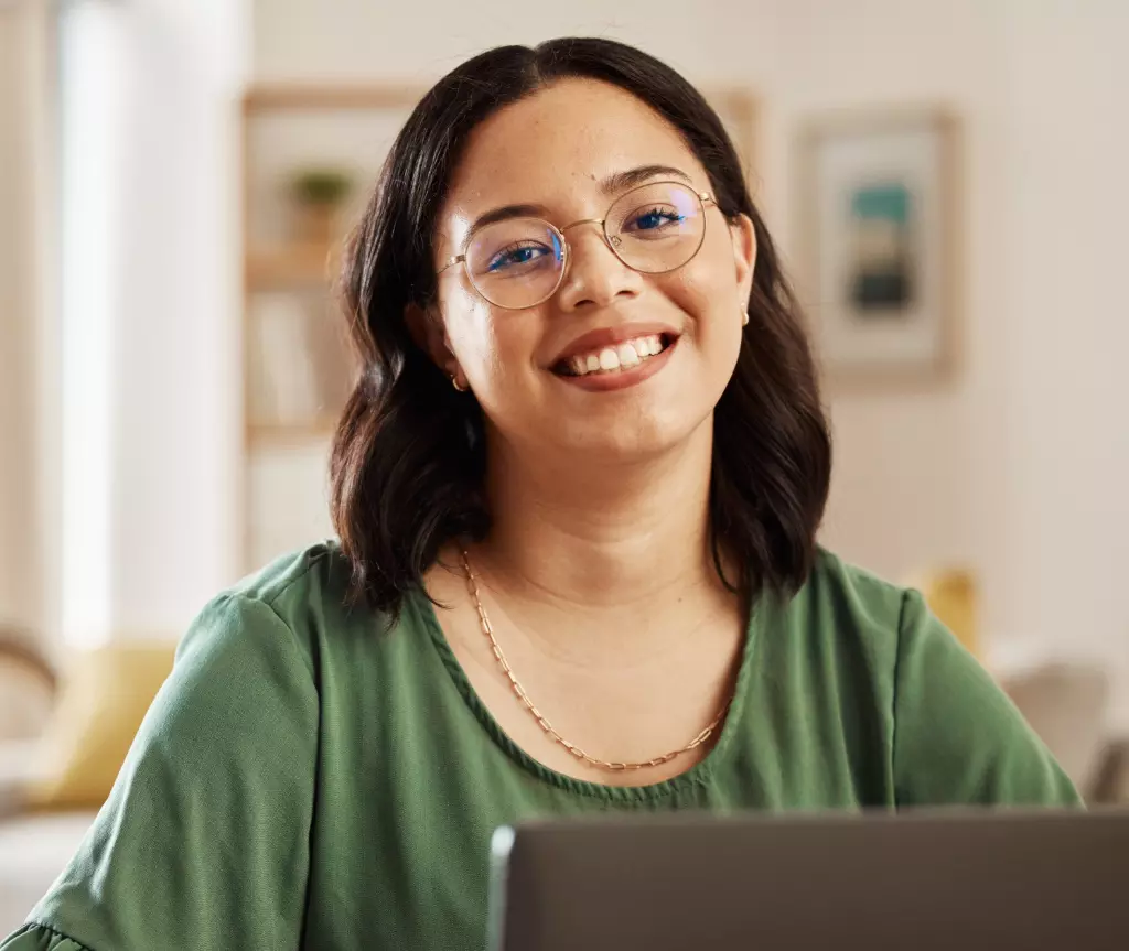 Una mujer joven con gafas, sonriendo, sentada frente a un portátil en una habitación bien iluminada, estudiando psicología.