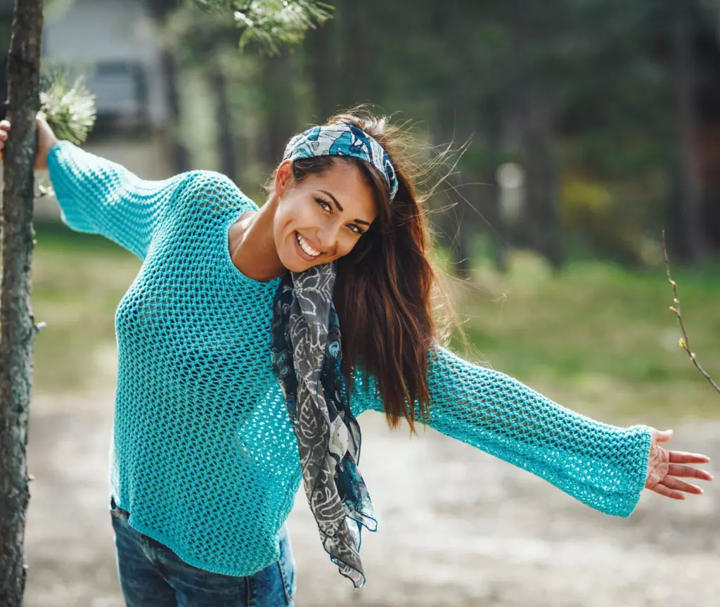 Una persona alegre con actitud de vida plena con un suéter de malla turquesa y una diadema que sonríe juguetonamente en un entorno forestal.