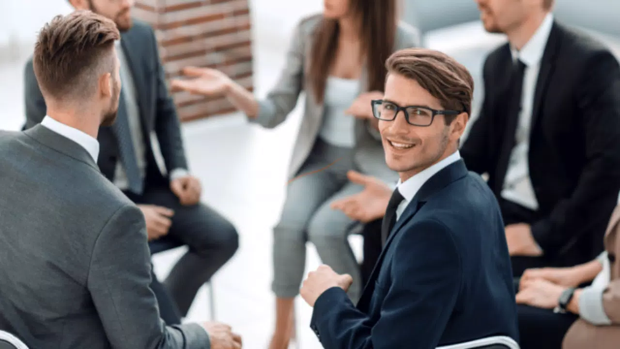 Un grupo de seis personas vestidas de negocios están conversando mientras están sentadas en círculo. Un hombre con gafas, que encarna la esencia del vivir con propósito, se gira hacia la cámara y sonríe.