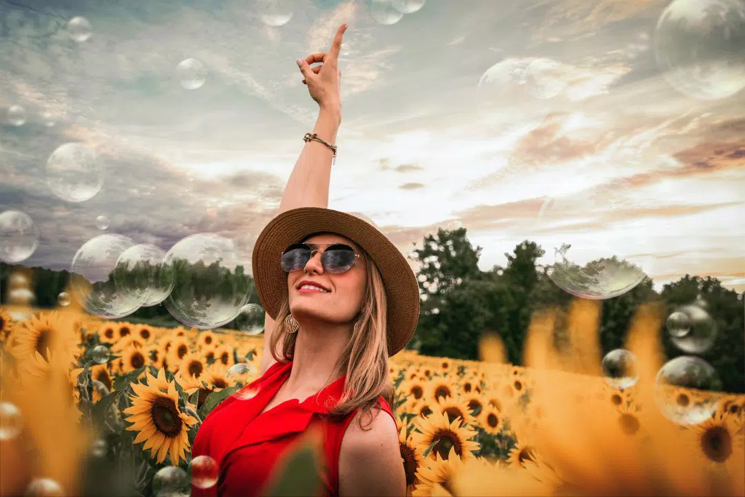 Mujer con un vestido rojo, sombrero y gafas de sol, parada en un campo de girasoles, señalando burbujas en el cielo con árboles al fondo: una escena perfecta que captura el bienestar y la tranquilidad.