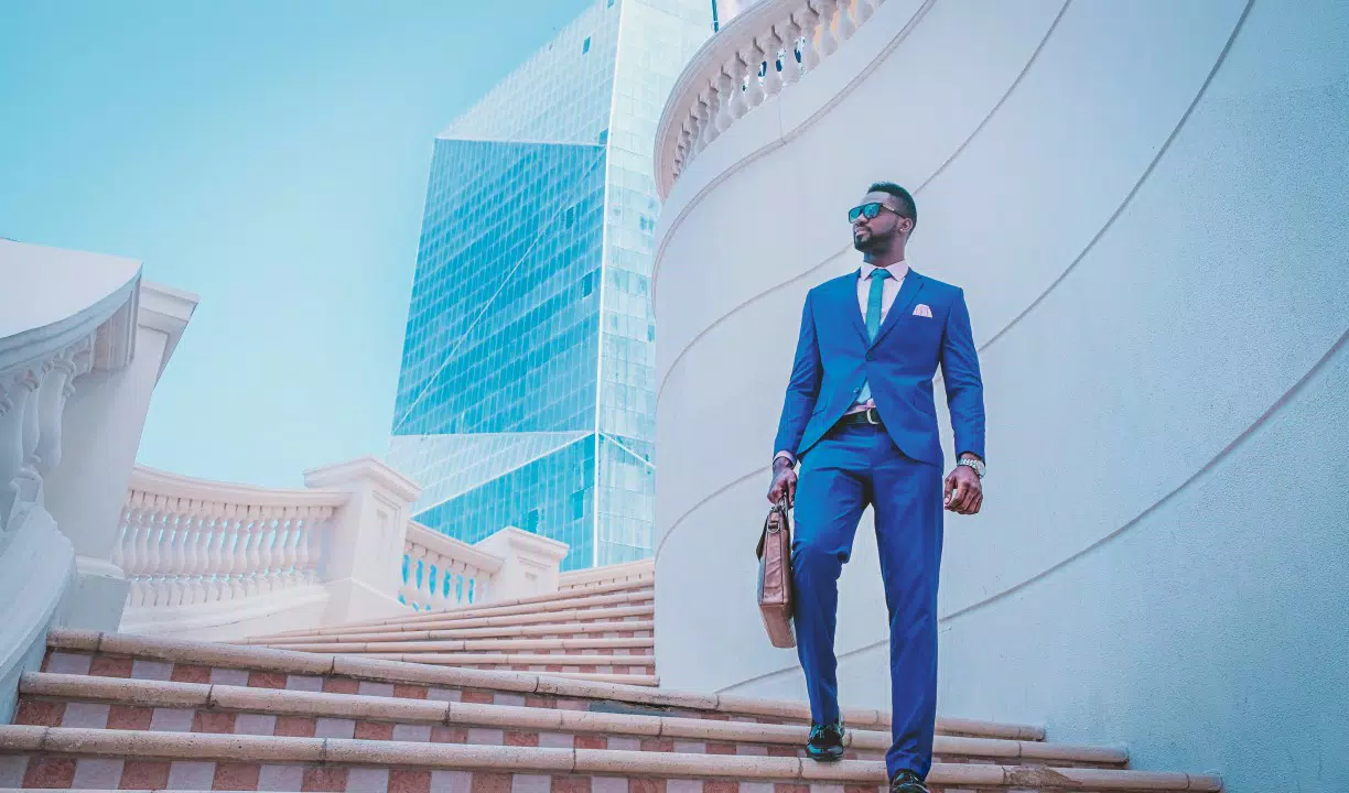 Un hombre con traje azul y gafas de sol, portando un maletín marrón, sube unas escaleras con modernos edificios de cristal al fondo, destilando un aire de bienestar.