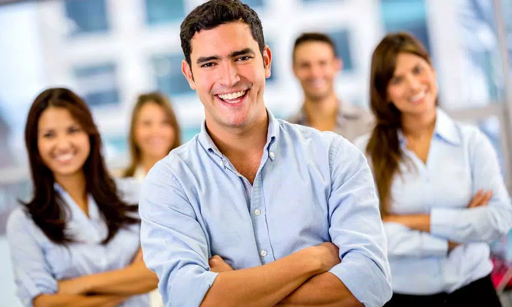 Un hombre con una camisa azul claro se encuentra frente a cuatro personas, sonriendo con los brazos cruzados, irradiando los principios de la psicología positiva. El fondo está borroso y todos parecen estar adentro.