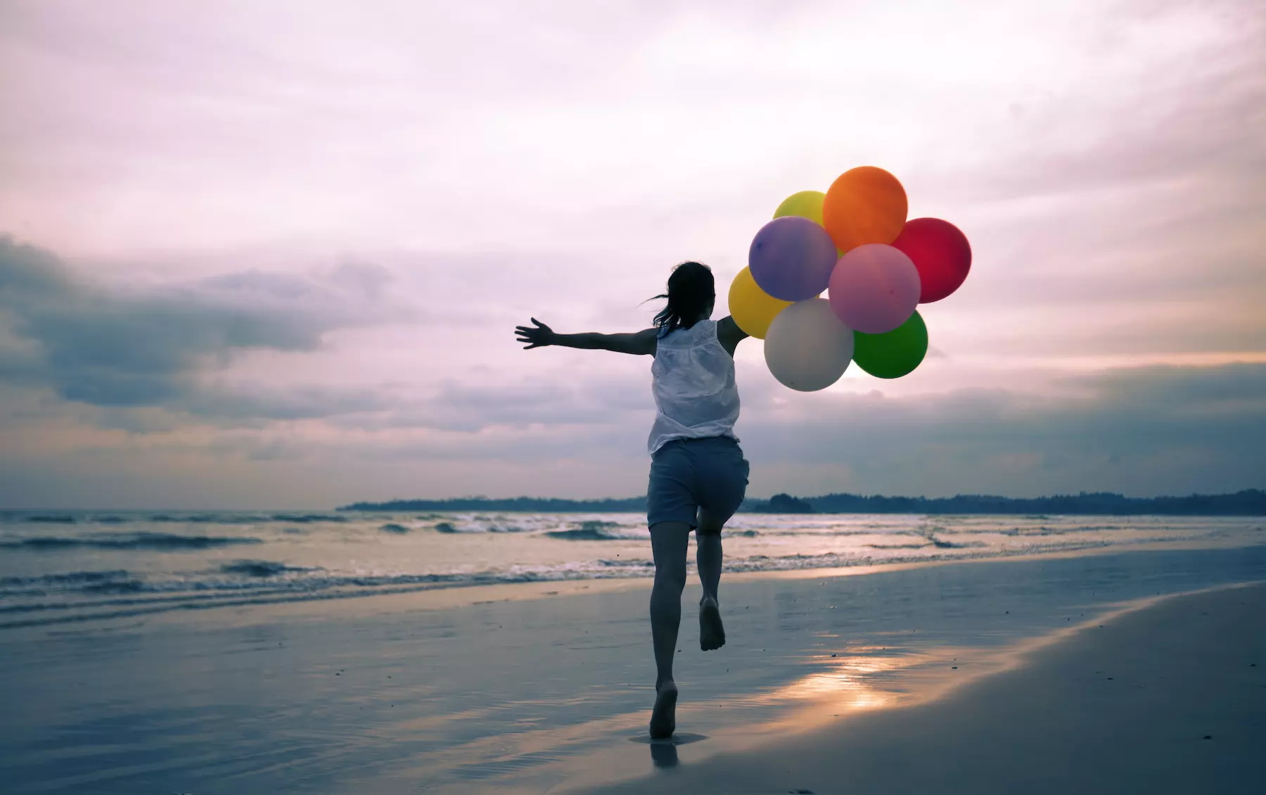 Una persona que sostiene globos de colores corre alegremente por una playa al atardecer, con el mar y el cielo nublado al fondo.