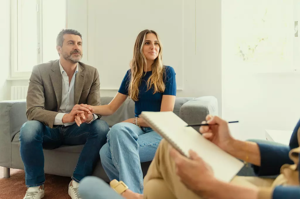 Una pareja en una sesión de terapia, sentada en un sofá, mirando atentamente, mientras un entrenador sostiene un portapapeles sentado en primer plano.