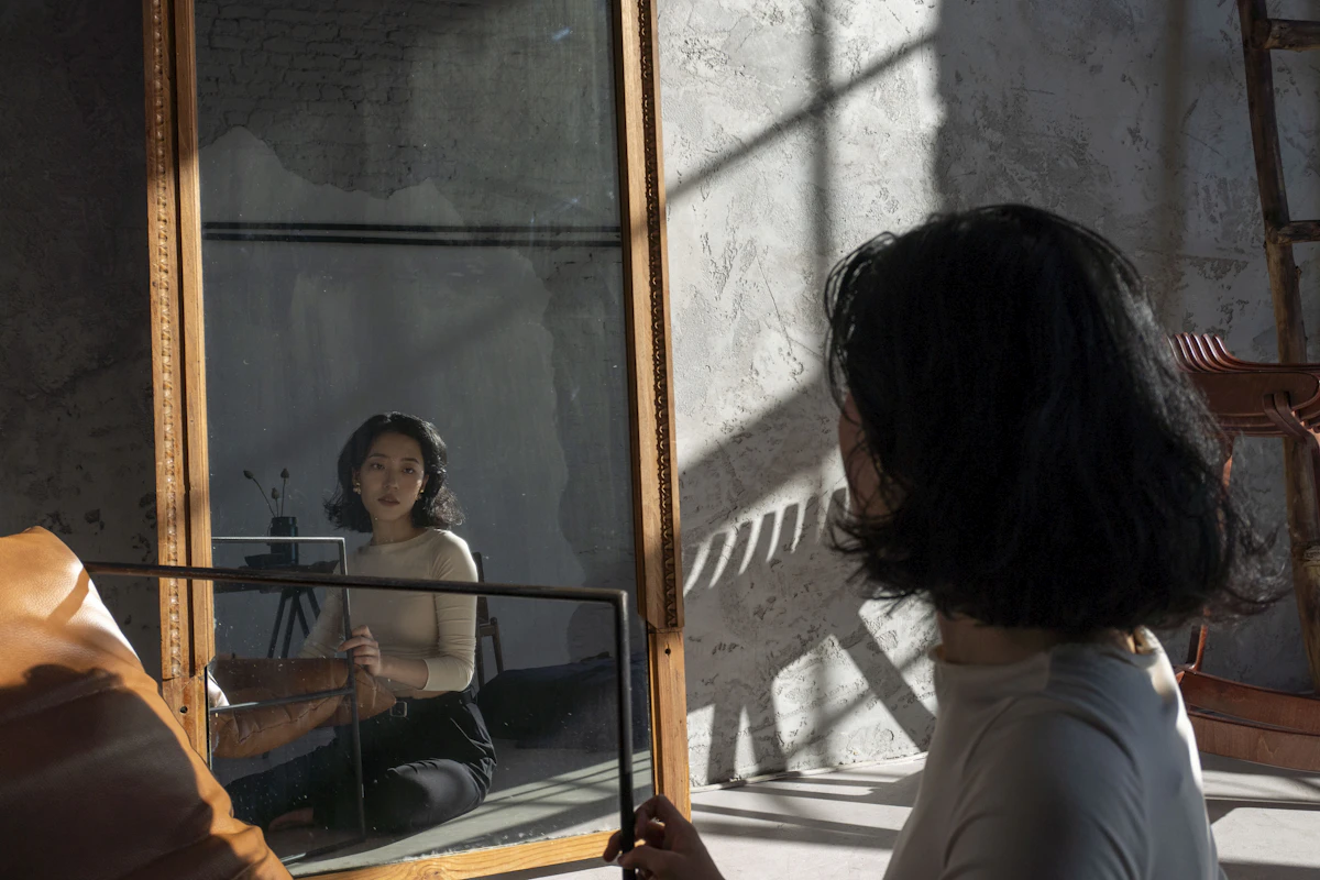 Una persona de pelo oscuro está sentada frente a un gran espejo enmarcado, en el que se reflejan su rostro y la parte superior de su cuerpo.