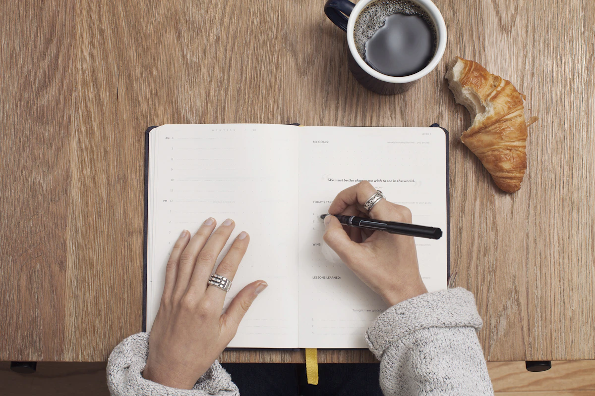Unas manos escribiendo en una agenda abierta sobre un escritorio de madera, junto a una taza de café y un cruasán.