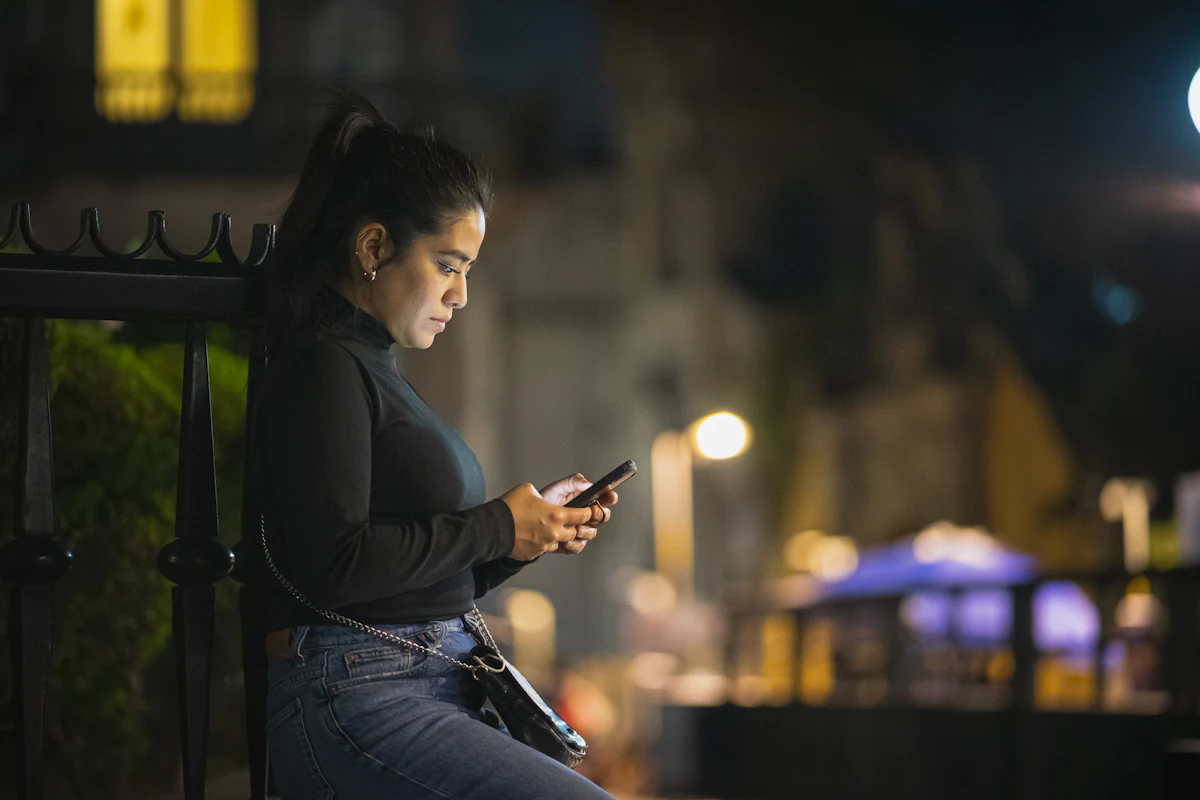 Una mujer apoyada en una valla por la noche, mirando su móvil.
