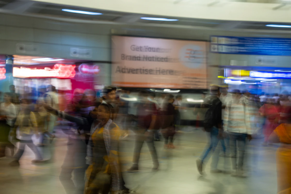 Multitud de viajeros, con efecto de movimiento difuminado, caminando por una estación de transporte público cubierta, con un cartel publicitario al fondo.