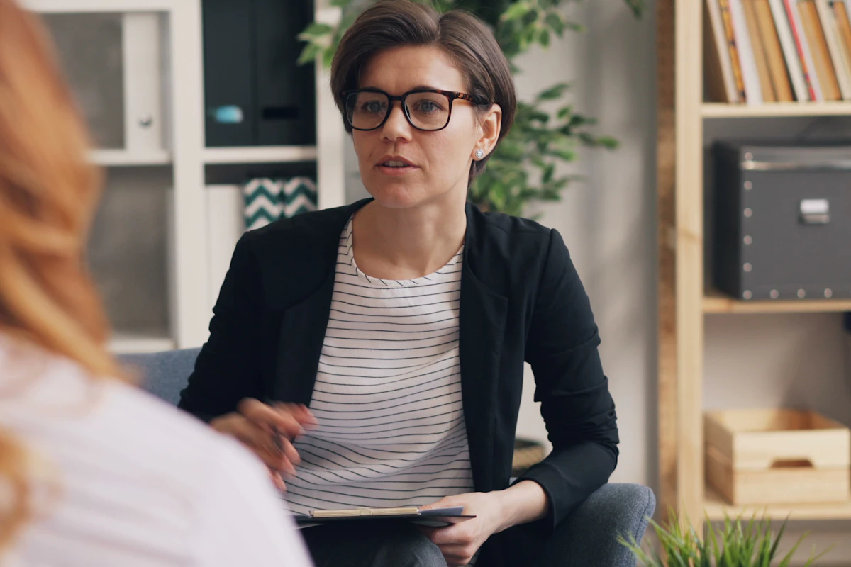 Una mujer profesional, con gafas y chaqueta, que sostiene un portapapeles, habla con un cliente en una oficina.